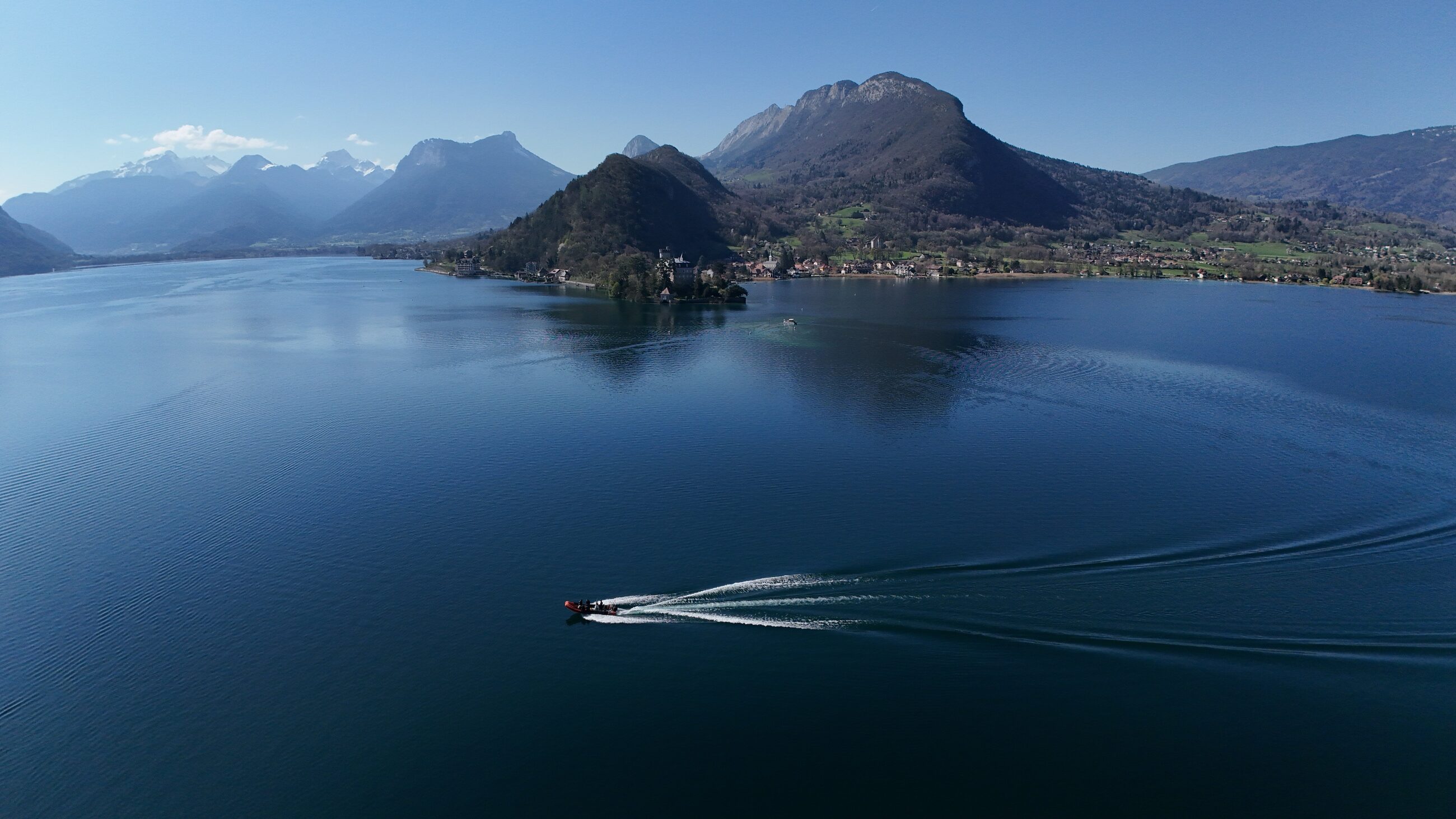 Aerial view of Lake Annecy, France — a boat cuts a wake across deep blue water below the Alps