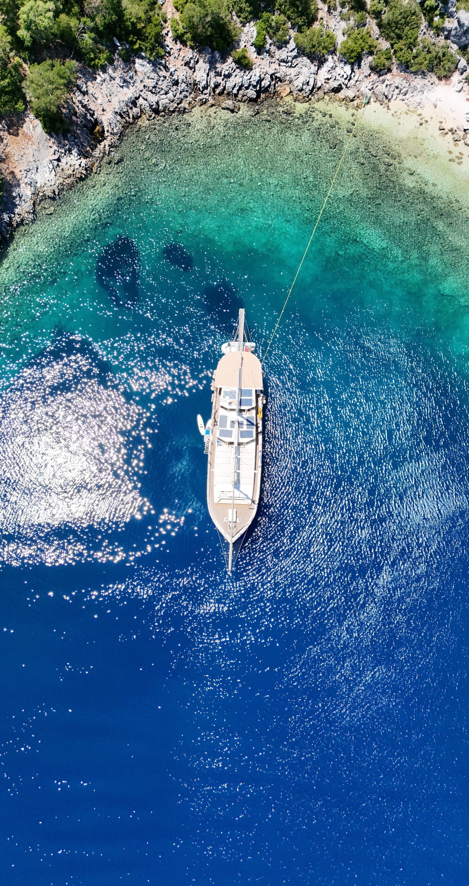 Top-down aerial of a gulet at anchor where deep blue water shelves up to a turquoise rocky shore