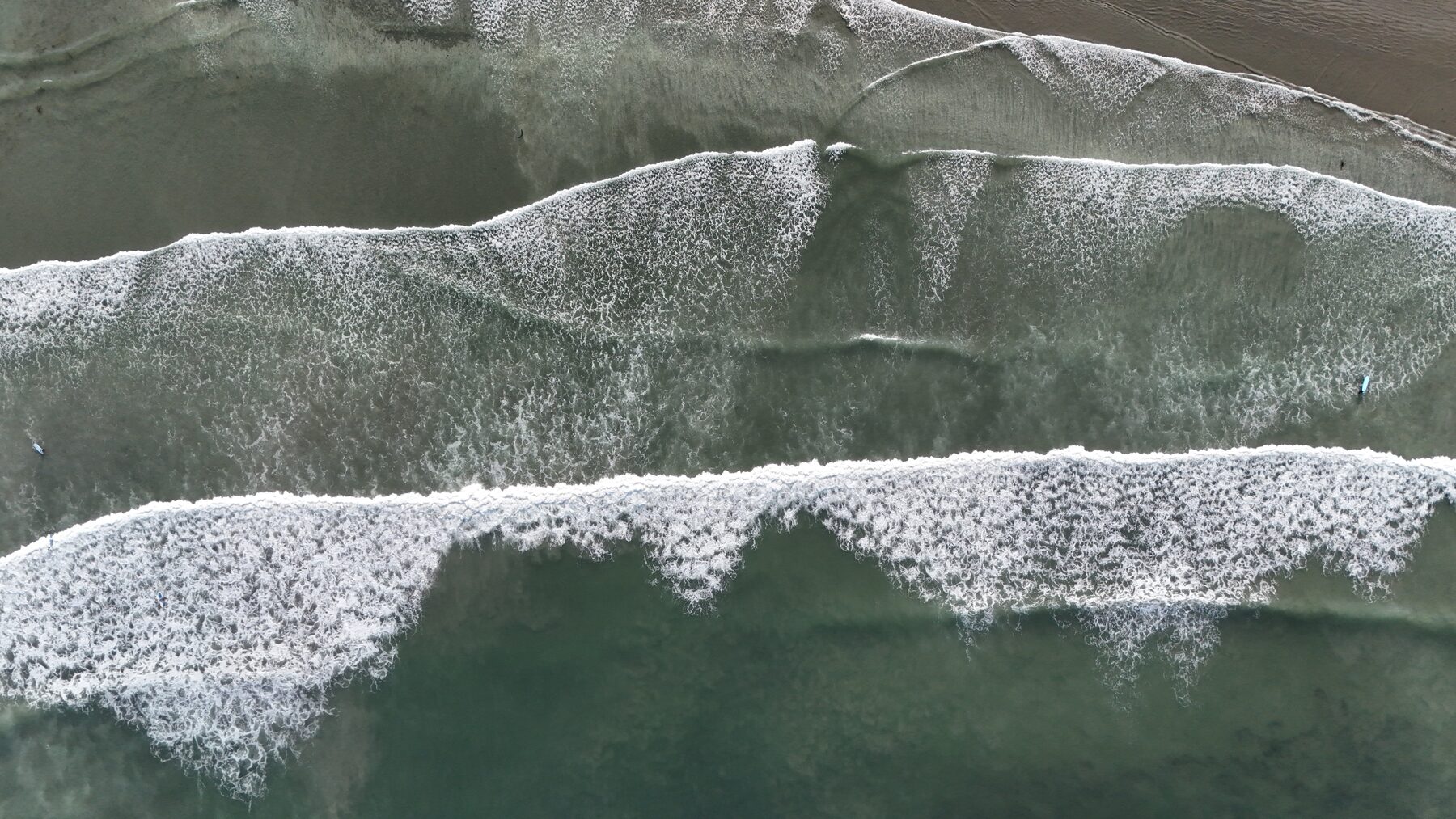 Top-down aerial of white water fanning across a sandy shorebreak