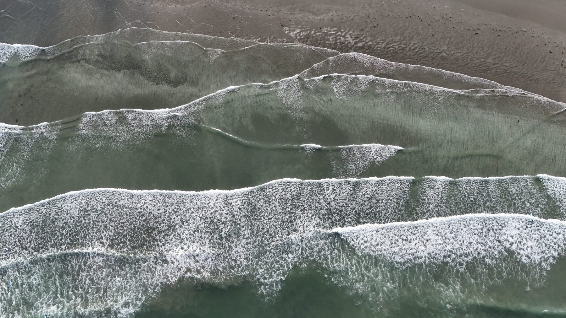 Top-down aerial of successive wave lines washing over tidal sand