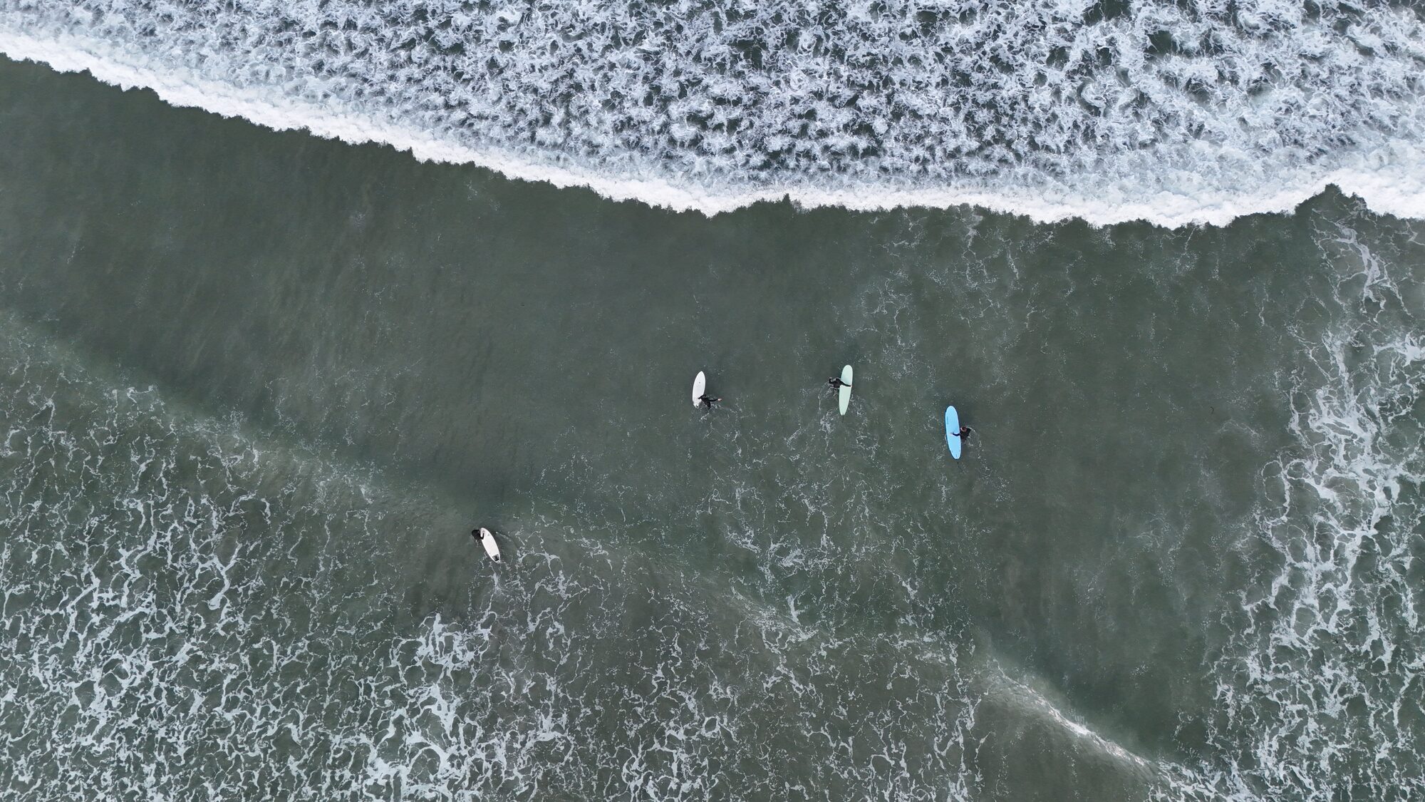 Top-down aerial of surfers waiting in the lineup beyond a breaking wave