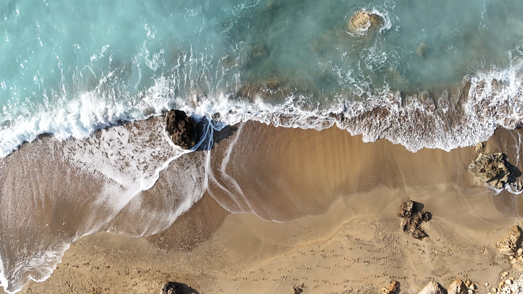 Top-down aerial of surf washing around dark rocks on golden sand