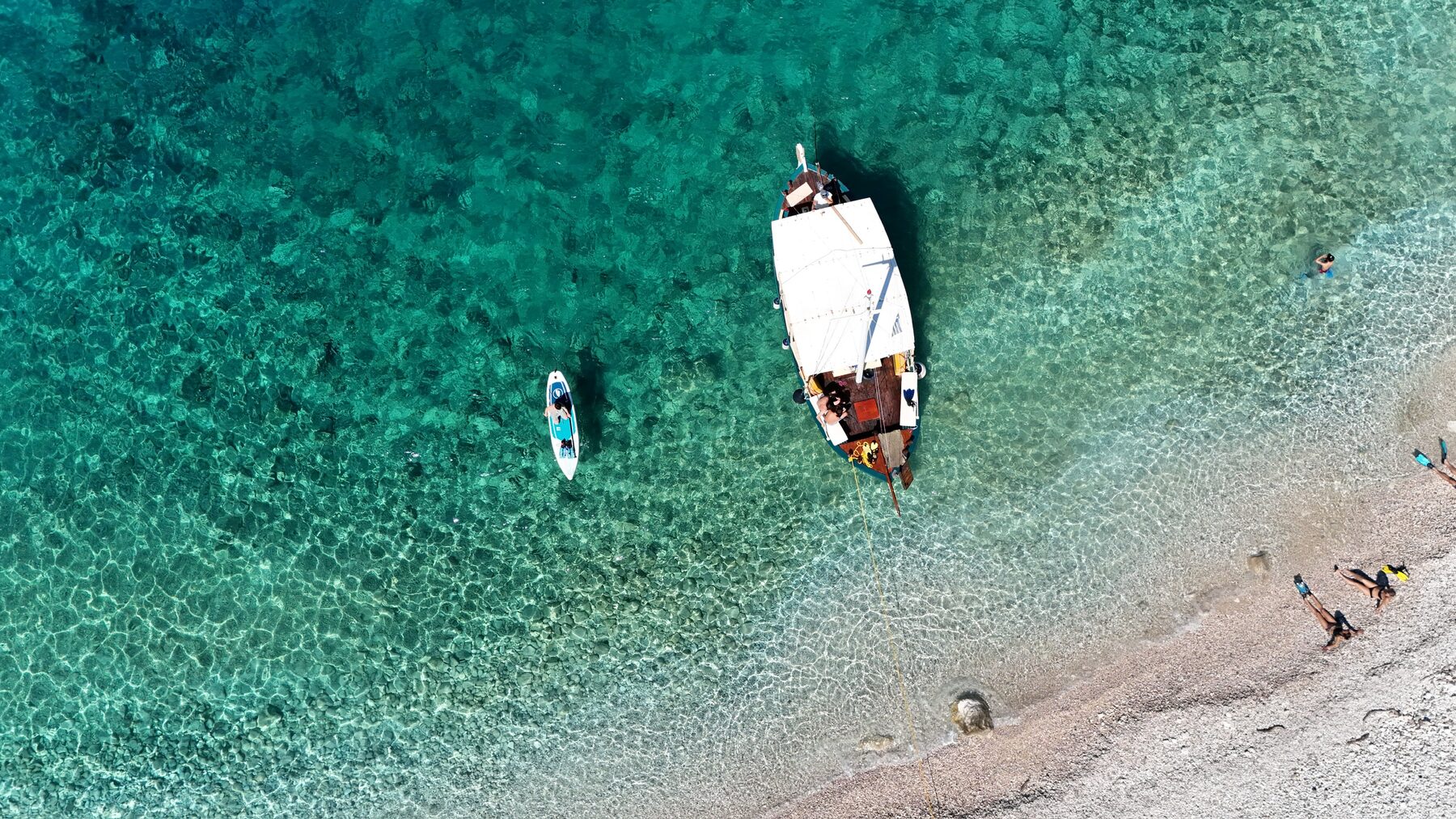 Top-down aerial of a traditional wooden boat moored in clear turquoise water off a pebble beach, with a paddleboarder and swimmers
